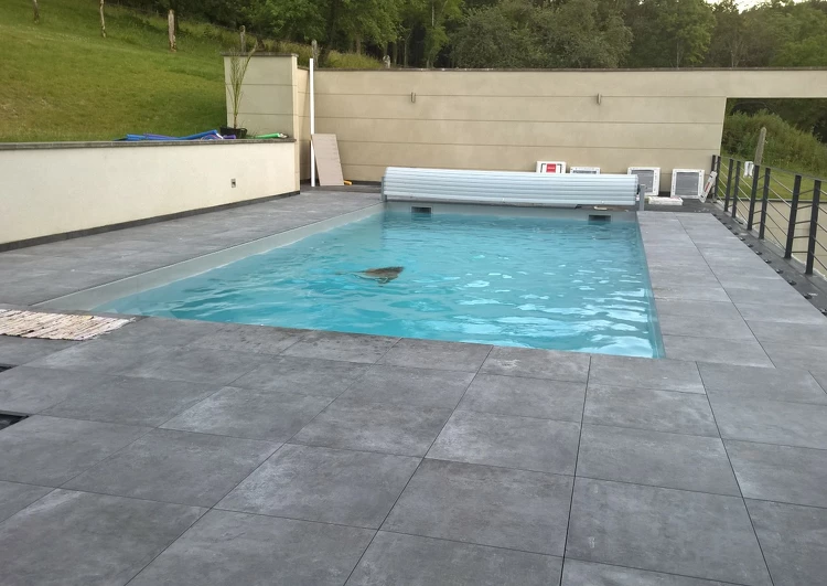 toiture terrasse avec une piscine, dalles sur plots, à gauche quelques dalles manquantes laisse voir la membrane EPDM, au fond un mur, à gauche des garde-corps, à l'arrière plan de la végétation.