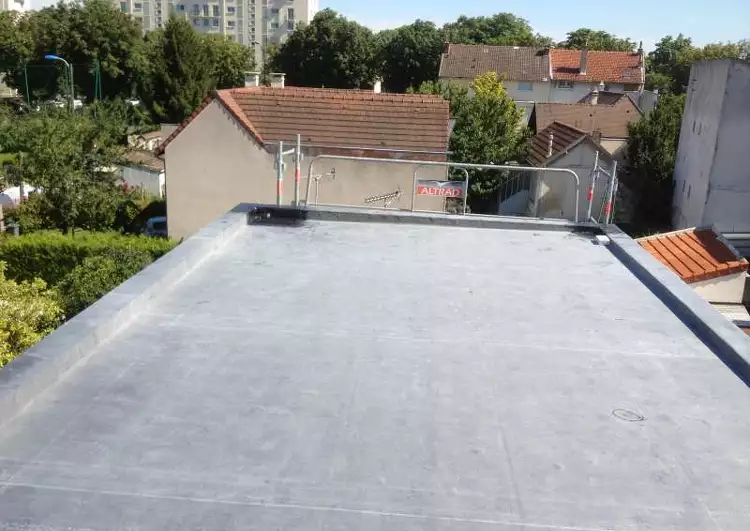 toiture plate bordée d'acrotères, couverte en membrane EPDM, vue prise en hauteur, à l'arrière-plan on voit d'autres maison, un immeuble, et de la végétation.
