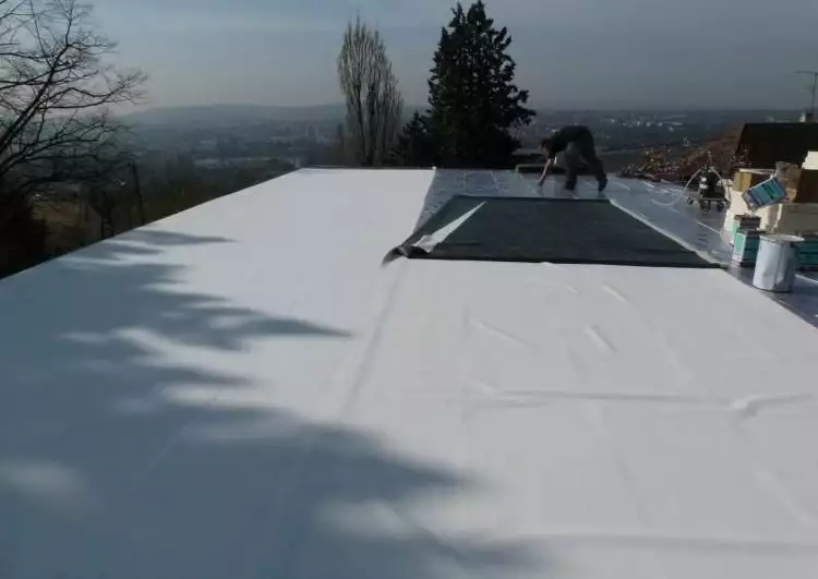 chantier en cours de pose de membrane EPDM blanche, sur la droite on voit des pots de colle et des outils, un ouvrier est en train d'appliquer la colle, l'horizon est visible à l'arrière-plan