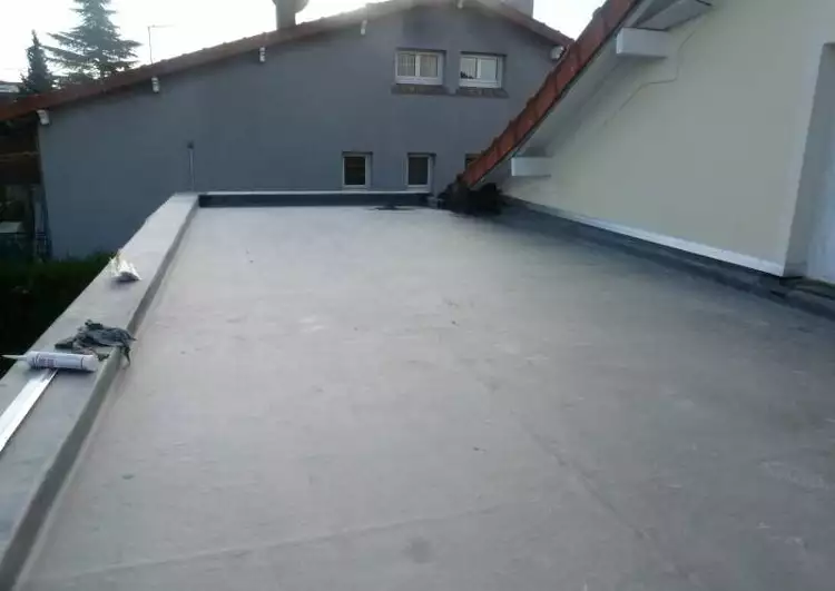 toiture plate couverte en membrane EPDM délimité à gauche et au fond par un acrotère, à droite par un mûr, des outils sont posés sur l'accrotère, à l'arrière-plan une maison.