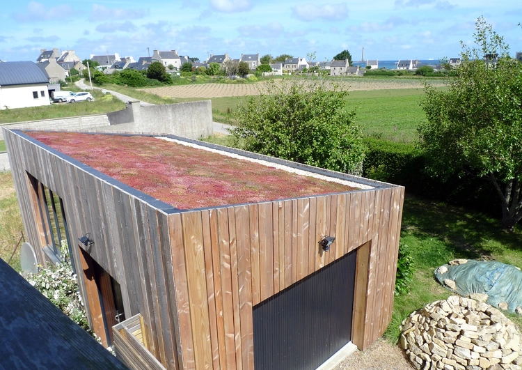 garage avec bardage en bois et toiture en pente douce avec une végétalisation extensive, à l'arrière plan un champ et un village.
