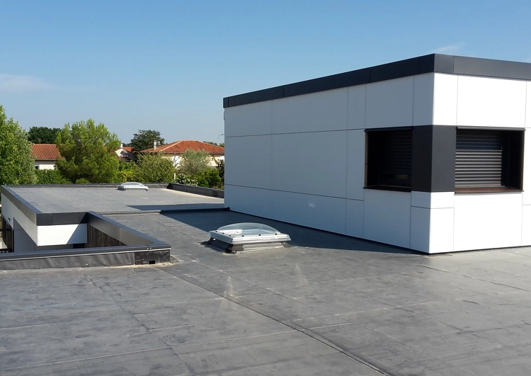 toiture plate au formes complexes couvertes en membrane EPDM, acrotères, puits de lumière, mur avec une fenêtre sur la gauche, à l'arrière plan une zone résidentielles avec des grands arbres.